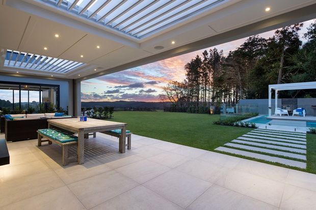 A contemporary covered outdoor entertaining area opens onto a landscaped lawn and pool terrace at dusk. The space features a large tiled patio, outdoor dining table, built-in seating, extensive glass sliding doors, and a translucent louvred/skylight roof structure. Beyond the lawn is a swimming pool with a white pavilion or cabana, while wooded hills and a sunset sky form the distant background.