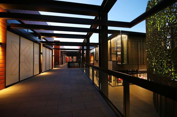 A covered elevated walkway or balcony runs through a modern multi-level building complex. Black steel framing forms a strong repetitive grid overhead and along the balustrade, with dark paved flooring, warm timber-clad walls, illuminated glass-fronted interiors, and a perforated or planted screen at the right. The space has a dramatic contemporary architectural character and appears to connect commercial or hospitality-style rooms.