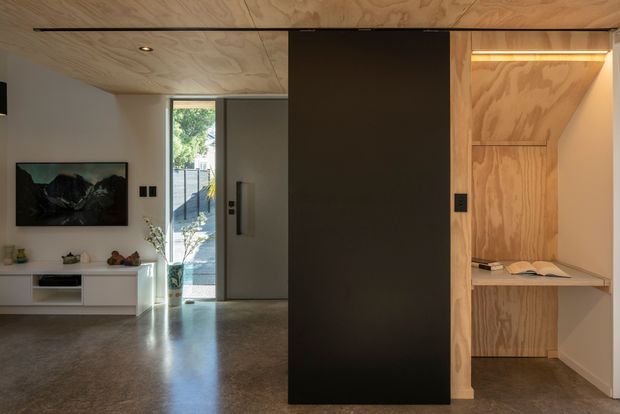 A compact contemporary interior featuring a built-in plywood-lined study nook or workspace beside a tall dark partition door. The room has a polished grey concrete floor, pale walls, timber plywood ceiling, recessed lighting, minimalist white low cabinetry, and a glazed entry opening to greenery outside. The timber-lined nook includes a slim desk shelf, open book, and integrated warm LED lighting.