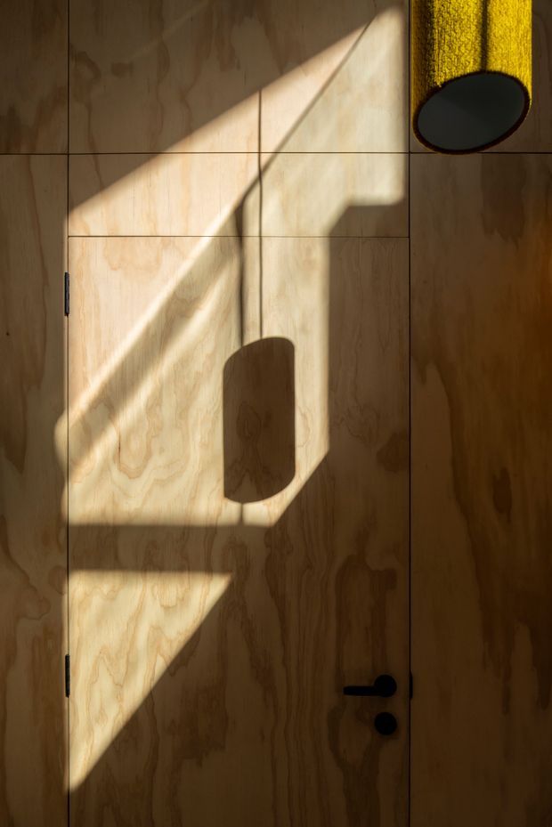 A close-up architectural detail of a plywood-lined interior wall and flush door, illuminated by strong geometric sunlight and shadows. The natural timber grain, dark door hardware, and a partial cylindrical yellow pendant light create a warm, minimalist composition.