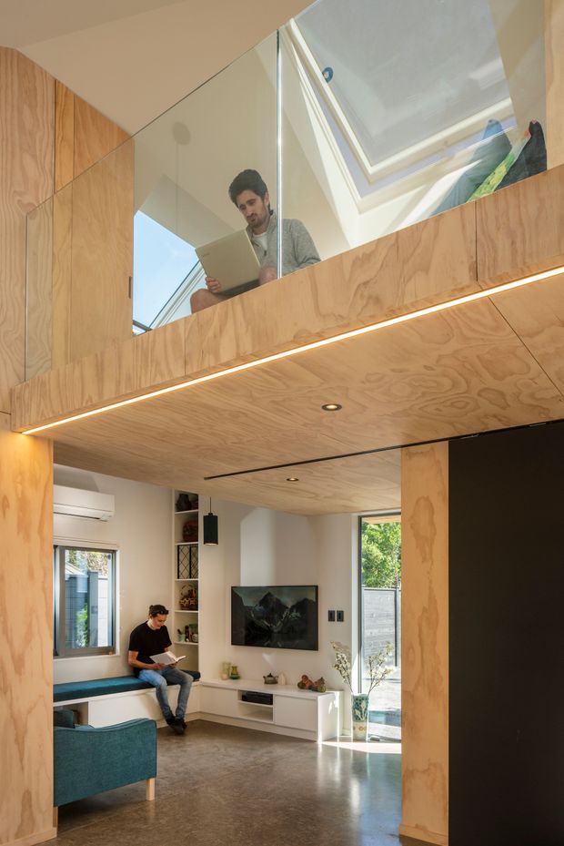 A compact, double-height contemporary living space with a mezzanine level and skylight. Plywood-lined ceilings, columns, cabinetry, and mezzanine balustrade create a warm timber envelope, while a glass balustrade overlooks the living room below. The lower level includes a built-in window seat, teal armchair, wall-mounted television, white media cabinetry, polished concrete floor, and large glazed doors opening to greenery. A person is seated with a laptop, and concealed linear lighting emphasizes the plywood ceiling edge.
