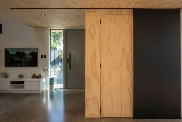 Interior view of a compact contemporary living and entry area. A full-height natural plywood-clad built-in volume with concealed doors dominates the foreground, contrasting with a matte black wall panel. The room has a plywood ceiling, polished concrete floor, white walls, recessed downlighting, a low white media cabinet, and a glazed entry door opening to the outdoors.