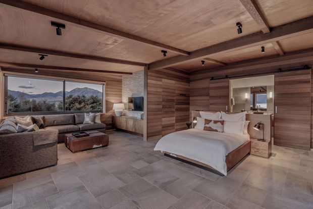 A warm, open-plan bedroom suite with a low-profile bed, bedside tables, lounge seating and a television area. The room features extensive timber wall and ceiling lining, exposed dark timber beams, large picture-window glazing with mountain views, and large-format grey floor tiles. A bathroom vanity is visible through the opening behind the bed.