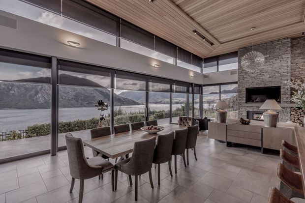 A refined open-plan dining and living area with floor-to-ceiling glazing overlooking a dramatic mountainous lake or valley landscape. A large rectangular dining table with upholstered chairs occupies the foreground, while a lounge area and wall-mounted television sit beside a full-height stacked-stone fireplace wall. The room features a slatted timber ceiling, dark window frames, pale large-format floor tiles, and contemporary built-in cabinetry.