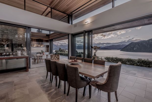 A sophisticated open-plan dining and kitchen area with a long timber dining table and upholstered dining chairs positioned beside expansive floor-to-ceiling glazing. The room features a dramatic timber-lined ceiling with exposed dark structural framing, dark-framed sliding or folding glass doors, pale large-format floor tiles, and a stone feature wall in the kitchen. Through the glazing is a striking mountainous lakeside landscape. The interior has a refined, warm contemporary character with strong indoor-outdoor connection.