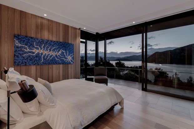 A refined bedroom with a large upholstered bed positioned beside a warm vertical timber-panelled feature wall. Floor-to-ceiling corner glazing provides expansive views over a lake and surrounding mountains. The room has pale timber flooring, light bedding, dark-framed windows, recessed ceiling lights, and a blue abstract wall artwork.