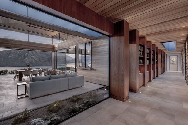 A refined contemporary interior hallway opens onto a spacious living and dining area through full-height glazing. Warm timber wall and ceiling lining, substantial timber columns, built-in shelving, pale stone or porcelain floor tiles, and a long skylit corridor define the space. The living area includes a large grey sectional sofa and dining table, while the glazing reveals a dramatic rocky landscape and water beyond.