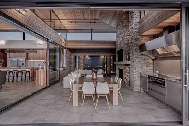 A refined open-plan indoor-outdoor entertaining space viewed through wide sliding glass doors. The room combines a dining area with a substantial stone-clad fireplace, an integrated stainless-steel outdoor-style kitchen, timber-lined ceilings and walls, and large-format grey flooring. A kitchen and breakfast bar are visible through the glazing on the left, while the central dining table opens toward a living area beyond.