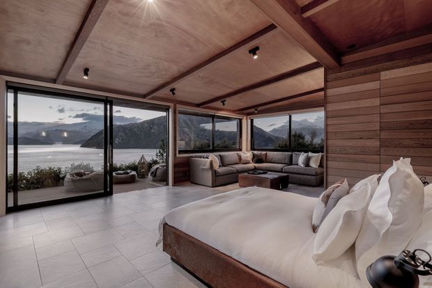 A spacious lakefront bedroom or retreat with a large bed in the foreground, a sectional seating area, and expansive black-framed glazing overlooking a mountainous lake landscape. The room features warm timber wall and ceiling panelling with exposed beams, pale tiled flooring, contemporary furnishings, and discreet track and recessed lighting.