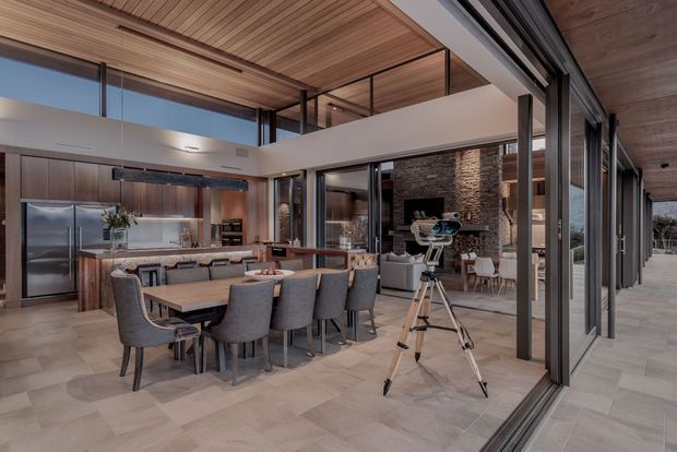 A spacious, open-plan contemporary interior combining a kitchen, dining area and living room. The room features full-height glazed sliding doors opening to an outdoor terrace, a high timber-lined ceiling with clerestory glazing, dark timber kitchen cabinetry, a long dining table with upholstered chairs, and a textured stone fireplace wall in the living area. A telescope stands near the glazing, with an outdoor view visible beyond.