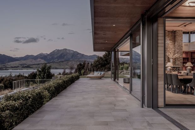 A contemporary lakeside residence viewed from a broad paved terrace, with full-height glazed sliding doors opening to a dining and living area. The terrace has a timber-lined soffit, dark framed glazing, and low hedging, with a lake and dramatic mountain range visible beyond.
