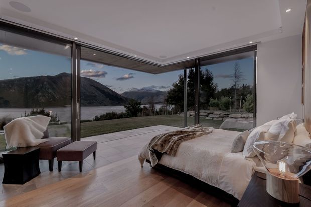 A contemporary bedroom with a low-profile bed, white bedding, warm wood flooring, and a pair of upholstered lounge chairs. Full-height corner glazing opens the room visually onto a paved terrace and landscaped grounds, with a dramatic mountain and lake view beyond. Recessed ceiling lighting and dark-framed sliding glass doors create a refined indoor-outdoor connection.