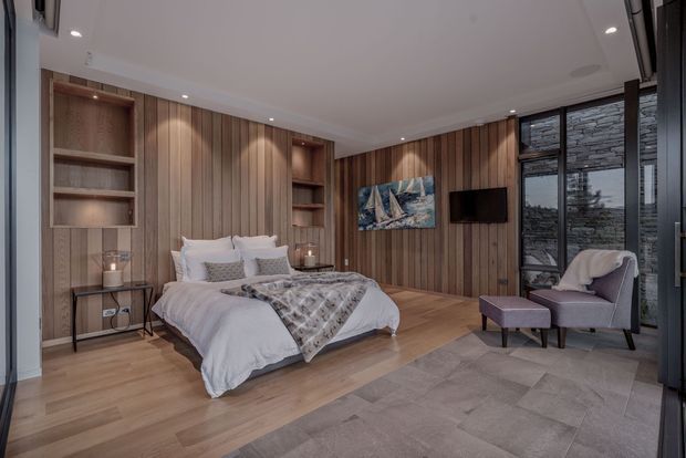A refined contemporary bedroom with a low-profile upholstered bed, white bedding, and integrated timber wall panelling with recessed shelving and warm accent lighting. The room combines timber flooring around the bed with large-format grey floor tiles in the sitting area. A small upholstered lounge chair and ottoman sit beside full-height dark-framed glazing, with a stone-clad exterior wall visible beyond.