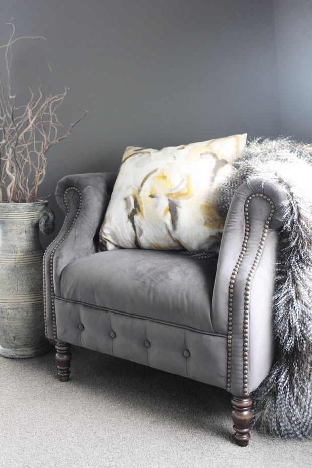A close-up interior vignette featuring a plush grey velvet wingback armchair with button detailing, rolled arms, turned wooden legs and metallic nailhead trim. The chair is styled with a cream, gold and grey decorative cushion and a draped grey faux-fur throw, set against a dark grey wall and neutral carpet. A tall aged metal planter with dried branches sits beside it.
