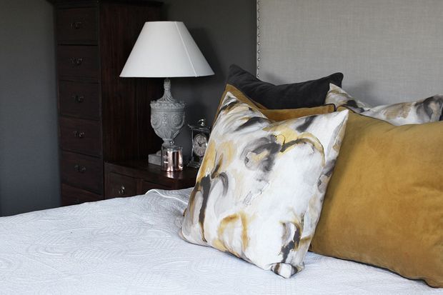A close-up of a styled bedroom featuring a neatly made bed with a white textured coverlet and layered decorative cushions in mustard, charcoal, cream, and abstract brushstroke patterns. A dark timber chest of drawers and bedside table sit against a deep grey wall, topped with an ornate white table lamp and small accessories.