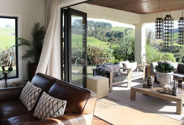 A bright, open-plan living room opening onto a covered outdoor terrace, with a brown leather sofa, neutral upholstered lounge chairs, timber coffee table, patterned cushions, large glazed openings, and distinctive black cage pendant lights. The room overlooks a lush rural landscape and is styled with natural materials and warm neutral tones.