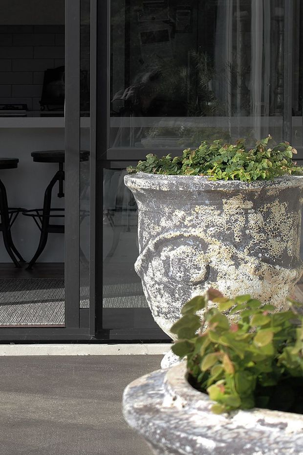 A close-up exterior view of weathered decorative concrete planters filled with greenery, positioned on a paved terrace in front of dark-framed glass doors. Through the glazing, a contemporary interior with a small bar-height table and stools is visible. The image emphasises the aged, textured planter surfaces, black window frames, and indoor-outdoor connection.