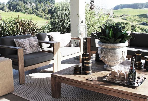 A bright covered outdoor terrace or veranda furnished as a relaxed lounge and entertaining area. Timber-framed armchairs and a large timber coffee table sit on a neutral outdoor rug, with a large planted urn, lantern, candles, glassware and champagne creating a refined outdoor setting. Rolling green rural landscape is visible beyond the terrace.