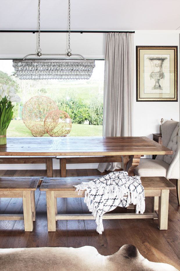 Bright dining room with a large rustic timber dining table and matching benches, positioned beside floor-to-ceiling glazing overlooking a garden. A crystal pendant chandelier, full-height taupe curtains, framed artwork, upholstered dining chair, and dark timber flooring create a refined rustic interior.