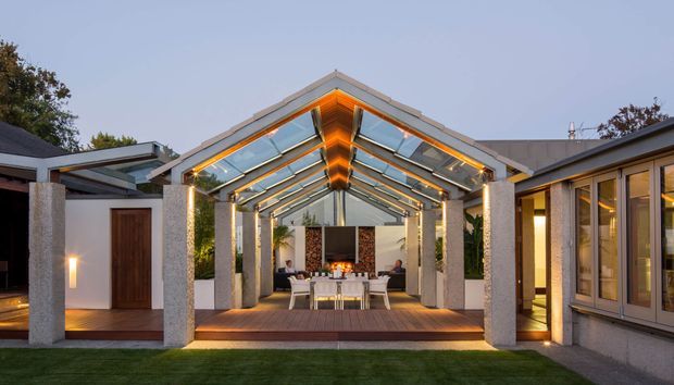 A dusk view of a sophisticated covered outdoor entertaining pavilion connected to a modern home. The pavilion has a pitched glazed roof with exposed timber beams, warm integrated lighting, textured light-coloured structural columns, and a timber deck. A dining table and seating sit around a central outdoor fireplace or fire feature, with glazed and solid building wings framing the space.