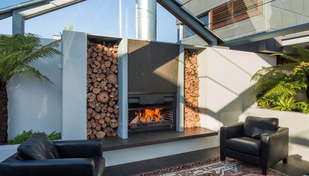 A contemporary outdoor lounge courtyard featuring a built-in open fireplace with a dark stone or concrete surround, flanked by recessed niches filled with stacked log rounds. Black leather lounge chairs sit on a patterned rug, while lush ferns and glass-lined modern architecture frame the space.