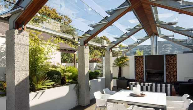 A bright contemporary outdoor dining and entertaining area beneath a glazed, angular canopy. The space features exposed timber beams, textured concrete columns, white rendered planter walls, lush planting, a white dining table with modern chairs, and a built-in dark outdoor fireplace or barbecue wall with stacked firewood.