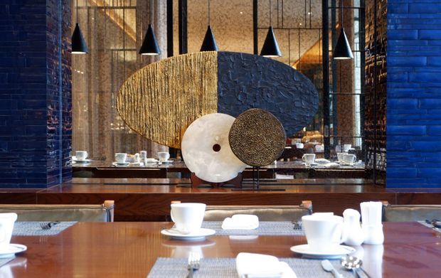 A refined restaurant dining area with polished wooden tables, white tableware, woven placemats, and upholstered banquette seating. The space features dramatic black cone pendant lights, a large sculptural circular artwork or screen, reflective surfaces, dark textured wall finishes, and tall patterned glazing, creating an elegant contemporary atmosphere.