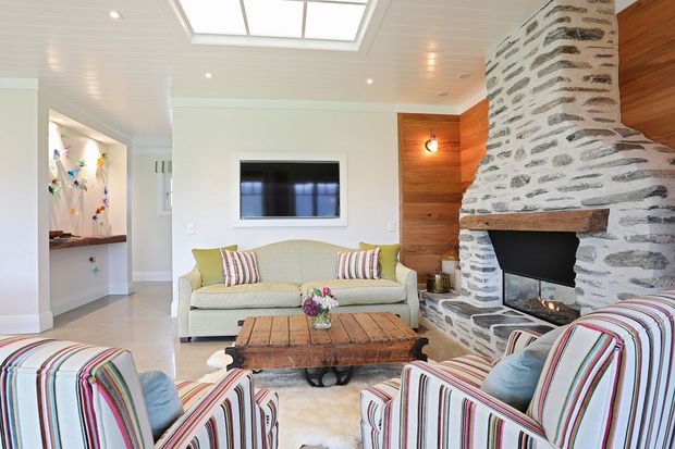 Bright, inviting living room with a large rustic stone fireplace, white panelled ceiling with skylight, warm timber wall panelling, built-in television, pale sofa, striped upholstered armchairs, and a reclaimed timber coffee table. The space combines coastal cottage comfort with rustic architectural detailing.