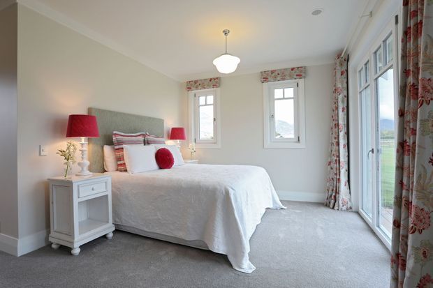 Bright, traditional-style bedroom with a neatly made bed, upholstered grey headboard, white quilt, red accent cushions and matching red bedside lamps. The room has pale walls, grey carpet, floral patterned curtains, multiple windows and full-height glazed doors opening to an exterior view.