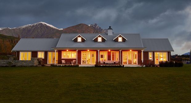 A long, single-storey countryside house photographed at dusk, with a symmetrical central gabled section, broad grey pitched roof, dormer windows, warm illuminated windows and doors, and stonework along the lower facade. A wide lawn fronts the home, with dramatic rugged mountains visible behind it.