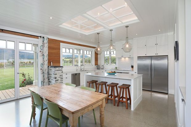 Bright open-plan kitchen and dining area with white cabinetry, a large central island, stainless-steel appliances, pendant lights, and a rustic timber dining table. Timber wall panelling, a tiled splashback, high windows, and a glazed roof opening bring warmth and natural light into the space, with views toward a green rural landscape.