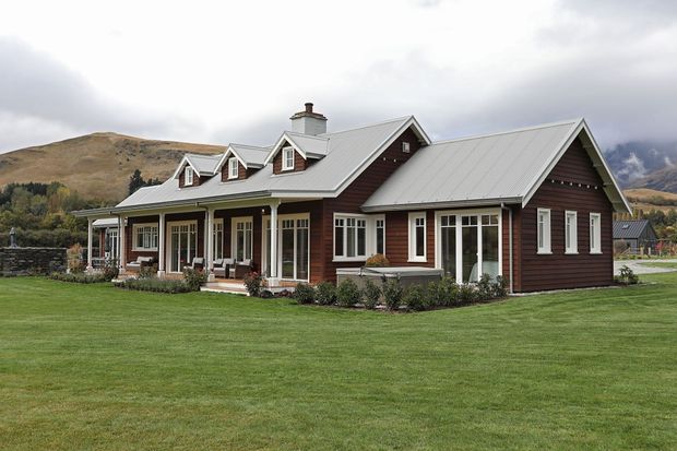 A large single-storey rural home with dark horizontal timber cladding, white trim, and multiple steep gabled roofs. The front façade includes a covered veranda, dormer windows, French doors, and many glazed openings. It sits on a broad manicured lawn with stone boundary walls and rugged mountain hills in the background under an overcast sky.