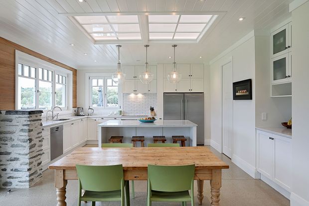 Bright, spacious white kitchen with shaker-style cabinetry, a central island, stainless-steel refrigerator, tiled backsplash, timber dining table, and green dining chairs. The room features a white beadboard ceiling with two large skylights, pendant lighting, extensive windows, and a rustic exposed stone breakfast-bar wall.