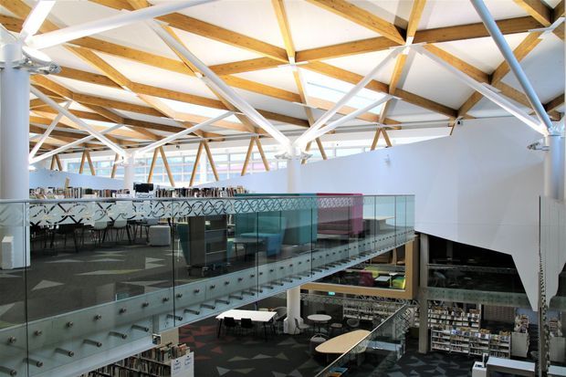 Interior view of a large contemporary multi-level commercial space, likely a library or hospitality venue, with a mezzanine, glass balustrades, extensive shelving and service counters. The dramatic exposed timber roof structure is painted and supported by white structural members, with abundant daylight and a high open ceiling.