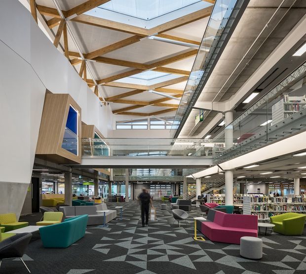 A spacious contemporary multi-level library or learning commons with open-plan seating, extensive bookshelves, glass balustrades, exposed timber roof trusses, skylights, concrete columns, and a geometric dark carpet. Brightly coloured lounge chairs and sofas create informal reading and collaboration areas.