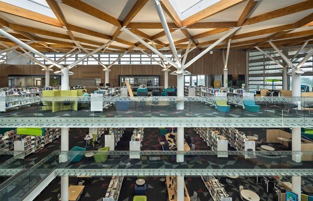 Wide interior view of a multi-level contemporary library or educational commercial space, showing extensive bookshelves, reading and study areas, glass balustrades, colourful seating, and a dramatic exposed timber-and-steel roof structure with high ceilings.
