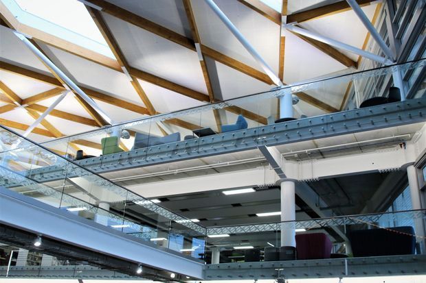 Interior view of a multi-level commercial or industrial-style building with exposed timber roof framing, white structural tubes and columns, steel beam edges, glass balustrades, and open mezzanine levels. The space appears bright and airy, with visible workspaces and furniture on the upper floors.