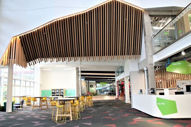 A bright contemporary commercial interior, likely a library, learning centre, or public information space. The open-plan area features a dramatic suspended timber-slat ceiling with a peaked form, white structural columns and walls, extensive glazing, yellow timber-and-metal tables and chairs, a white reception or information desk, and patterned dark carpet. Green-and-white 'Ask Me' signage is visible at the service counter.