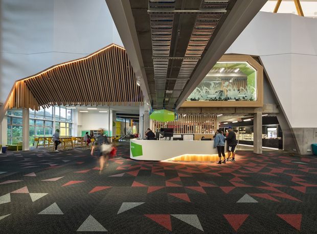 A large contemporary commercial interior, likely a public lobby or reception area, featuring a central white reception counter, exposed overhead services, dramatic angular timber slatted ceiling structures, extensive glazing, and a mezzanine-like timber-framed feature with artwork or planting behind glass. The patterned dark carpet incorporates scattered geometric red, grey, and beige shapes, while people and workstations occupy the open space.