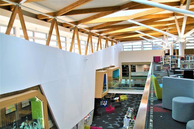 Bright contemporary library or learning space with an open mezzanine-like upper level, extensive bookshelves, colourful lounge seating, and a distinctive exposed timber and white-painted steel roof structure. Large windows and clerestory glazing provide abundant natural light, while geometric patterned carpet and integrated display joinery add visual interest.