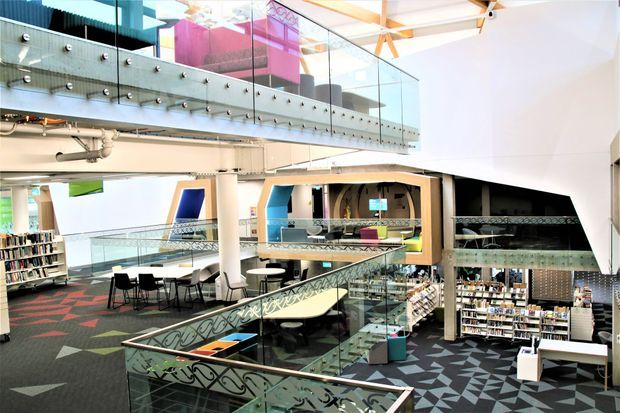 Interior view of a large multi-level library or commercial learning space with open mezzanine walkways, glass balustrades, exposed structural and service elements, bookshelves, study tables, and boldly patterned geometric carpet. The space has a bright, contemporary fit-out with colourful graphic accents and high ceilings.