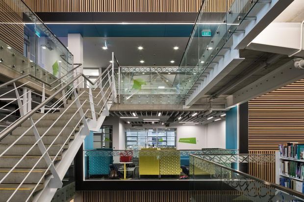 A contemporary multi-level commercial interior, likely a library or educational workspace, featuring an open stairway, mezzanine walkways, extensive glass balustrades, exposed services, timber slatted wall and ceiling accents, and colourful built-in seating or study areas. Bookshelves are visible on the right.