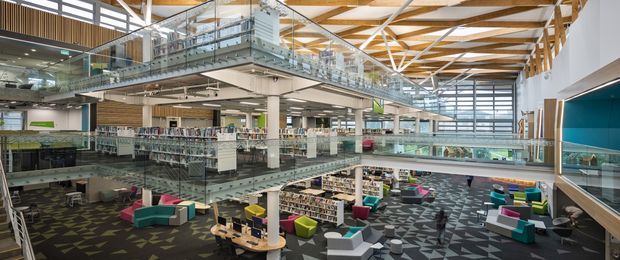 Large multi-level commercial library or civic interior with open-plan reading and lounge areas, extensive bookshelves, glass balustrades, exposed timber roof trusses, clerestory glazing, and colourful contemporary seating. The space has a high ceiling and a bright, expansive atrium-like layout.