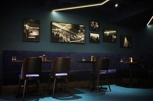 A moody, dark restaurant or hospitality interior with deep teal walls, a long upholstered banquette, small dining tables, tall dark dining chairs, framed monochrome artwork, and angular linear LED lighting across the ceiling. The space has a refined, intimate atmosphere with blue-toned lighting and dark finishes.