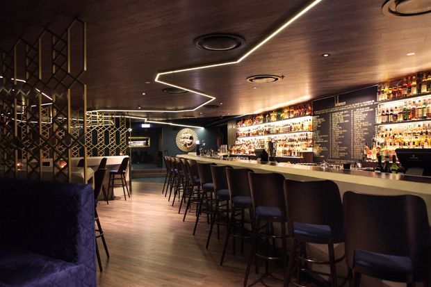 A moody, sophisticated bar interior with a long illuminated drinks counter, extensive backlit bottle displays, dark upholstered bar stools, timber-look flooring and a dark timber ceiling. Decorative geometric metal screens divide the seating area, while linear LED lighting and recessed fixtures create a dramatic atmosphere.