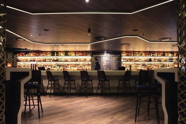 A sophisticated bar interior with a long illuminated liquor display, central service counter, and rows of dark bar stools. The space features a warm dark timber ceiling with recessed and linear lighting, wood-look flooring, and black structural lattice screens framing the sides.