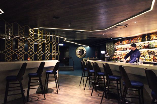 A stylish dark-toned cocktail bar interior with a long illuminated bar counter, extensive backlit liquor shelving, high stools, geometric metal screenwork, dark timber ceiling, and integrated linear lighting. A bartender is working behind the bar.