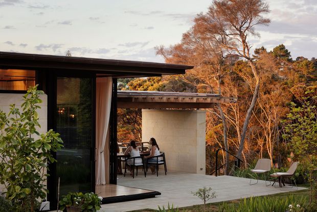 A modern single-level home opening onto a broad outdoor terrace in a wooded setting. The scene features dark-framed floor-to-ceiling glazing, warm timber interior lining, slender flat roof planes, a pale masonry outdoor wall, dining furniture, and lounge chairs. Autumn-toned trees surround the quiet, integrated indoor-outdoor living area.