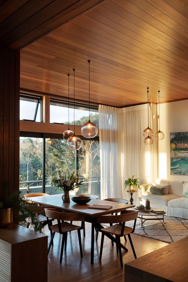 Warm, refined open-plan interior featuring a timber-lined vaulted ceiling and matching dark timber flooring and joinery. A dining table with six chairs occupies the foreground, while a light upholstered sofa and coffee table form a living area beside large windows with sheer curtains. Multiple spherical pendant lights hang over the dining zone, and abundant glazing provides views of trees outside.