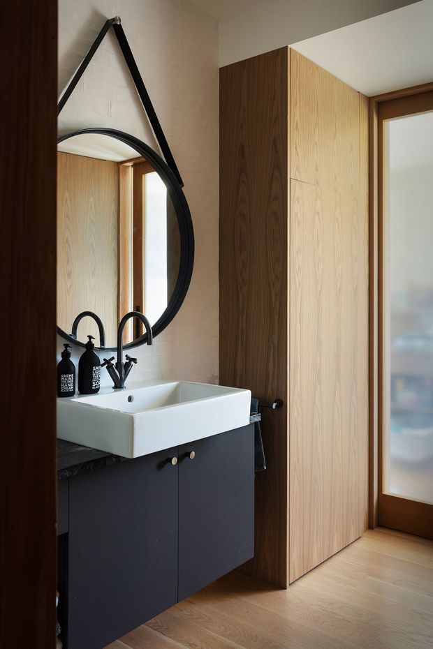 A compact, refined bathroom featuring a wall-mounted white rectangular basin on dark navy cabinetry, black curved tapware, and a large round black-framed mirror suspended by a leather strap. Warm timber built-in cabinetry, timber-framed frosted glazing, and light timber flooring create a calm, contemporary interior with strong tonal contrast.
